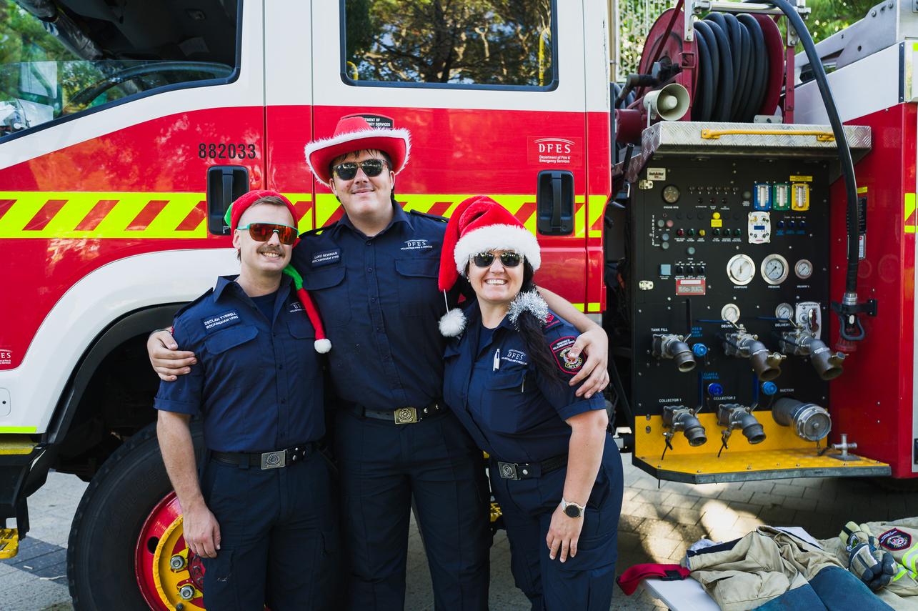 Three emergency services workers wearing Christmas hats smile in front of a fire truck.